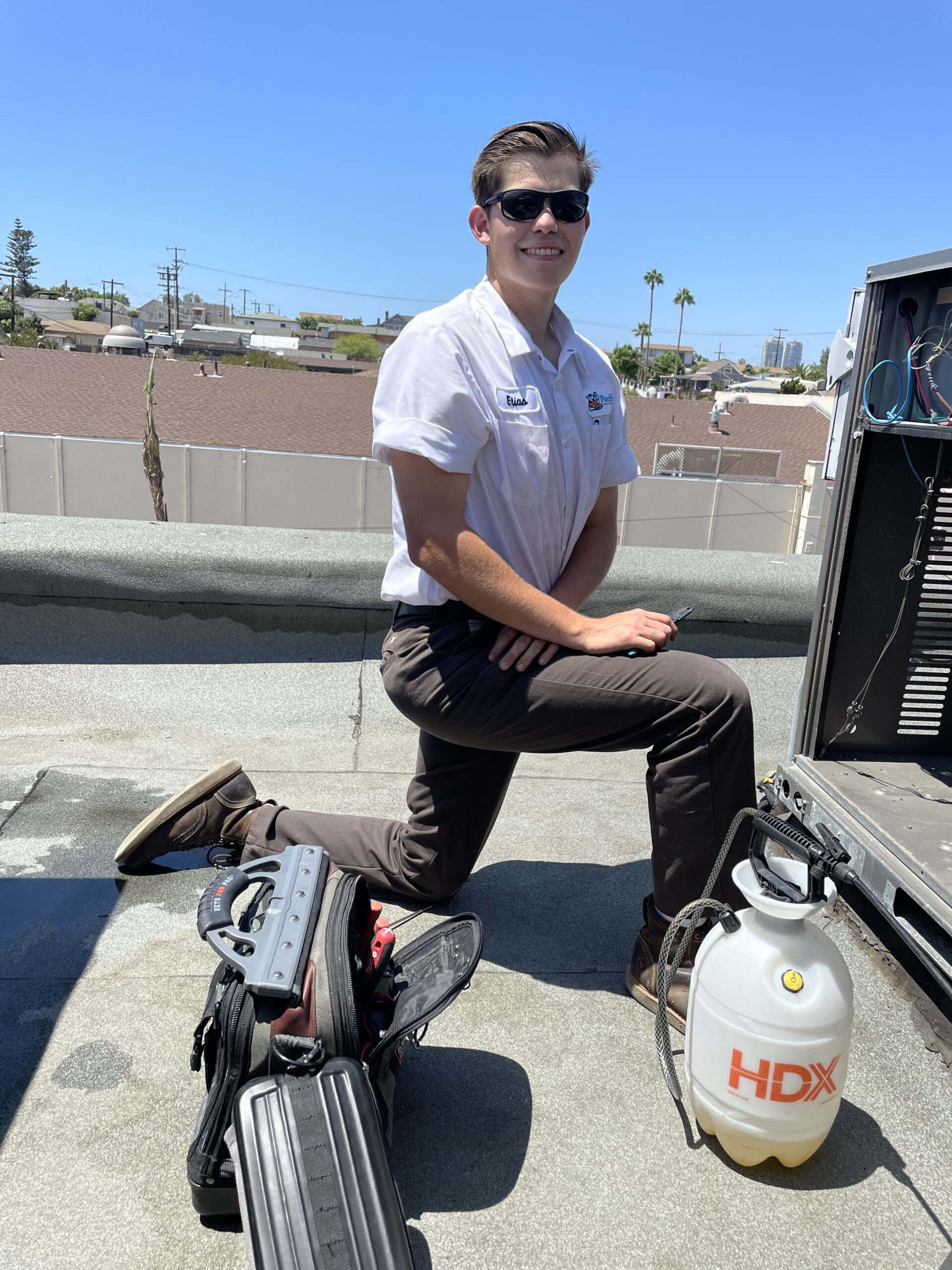 HVAC technician kneeling on a rooftop while servicing an air conditioning unit under a clear blue sky.